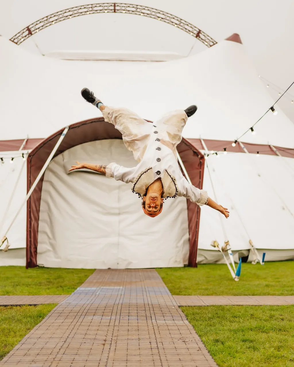A performer in white flips upside down on the path outside The Revel Puck Circus big top